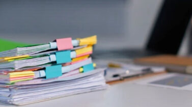 Stack of colorful, binder-clipped documents on a desk beside a laptop, notebook, and stationery.