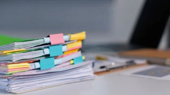 Stack of colorful, binder-clipped documents on a desk beside a laptop, notebook, and stationery.