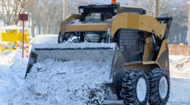 Yellow skid-steer loader clearing snow from a road on a sunny winter day.