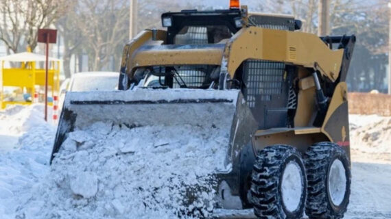 Yellow skid-steer loader clearing snow from a road on a sunny winter day.