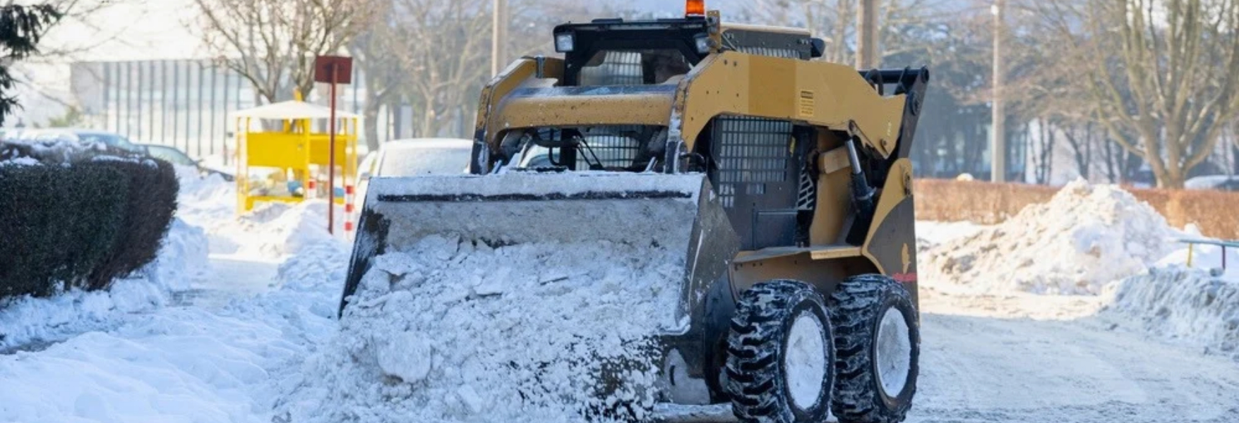 Yellow skid-steer loader clearing snow from a road on a sunny winter day.